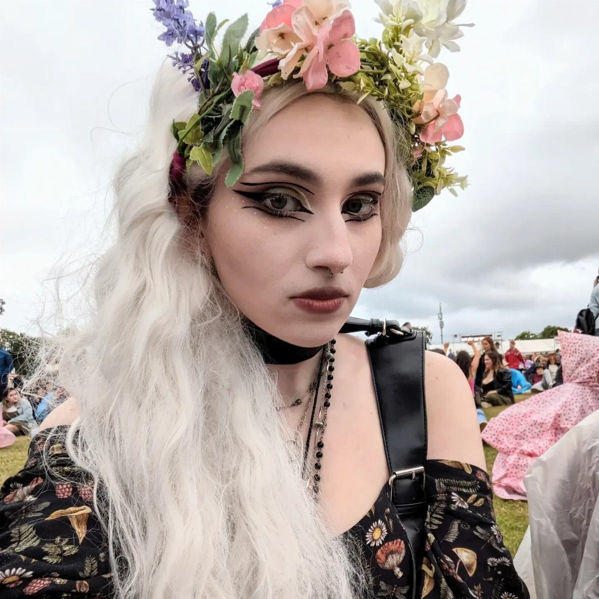 A woman with white hair wearing a flower crown and dark floral clothing outdoors.