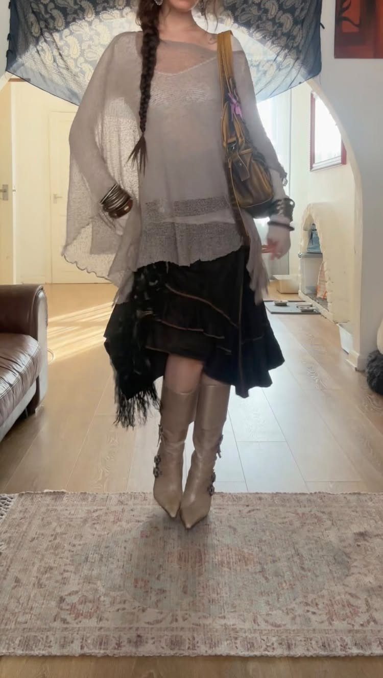 A person wearing a sheer grey top, black ruffled skirt, and tan boots standing indoors.