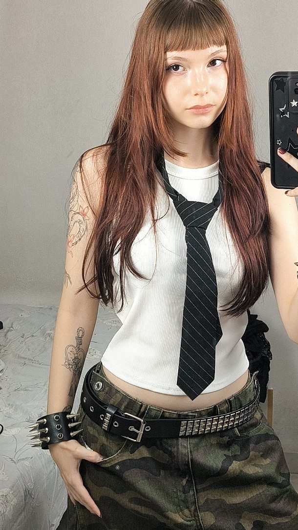 Young woman posing in a white tank top, black tie, and camouflage pants.