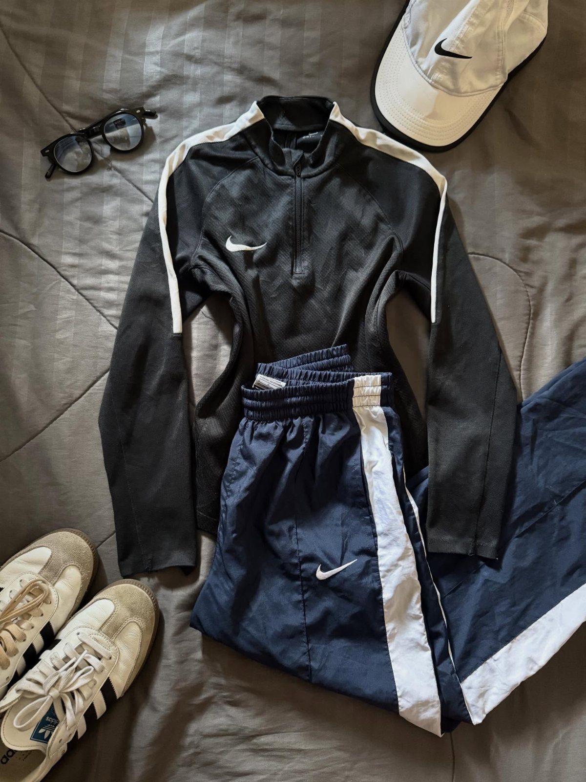 Nike athletic apparel including a black quarter-zip, navy track pants, white sneakers, and a baseball cap.