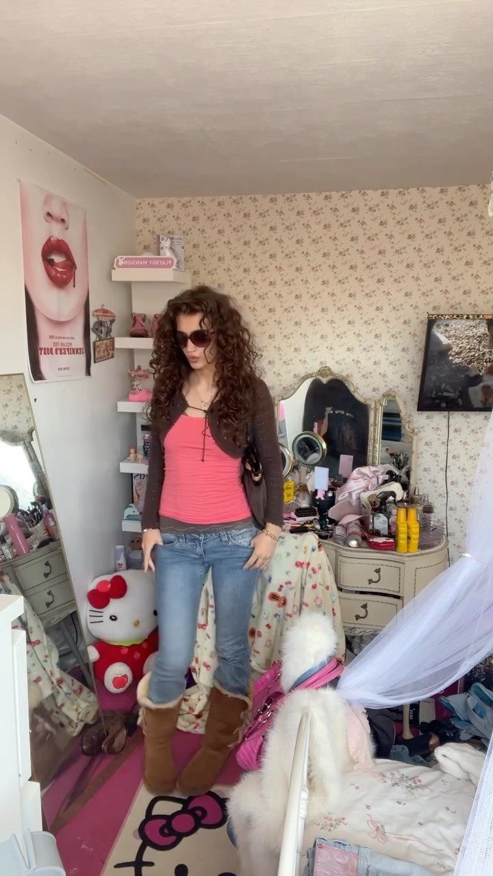 A woman posing in a bedroom wearing a pink tank top, brown cardigan, and blue jeans.