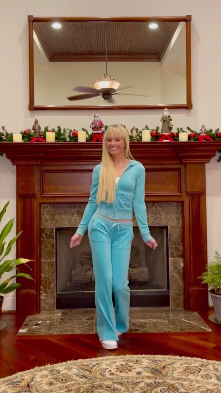 Blonde woman posing in a light blue velour tracksuit in front of a fireplace.