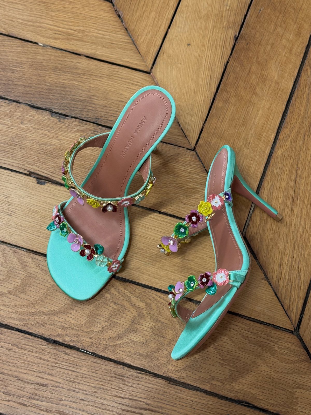 Mint green high-heeled sandals with colorful 3D floral decorations resting on a wooden floor.