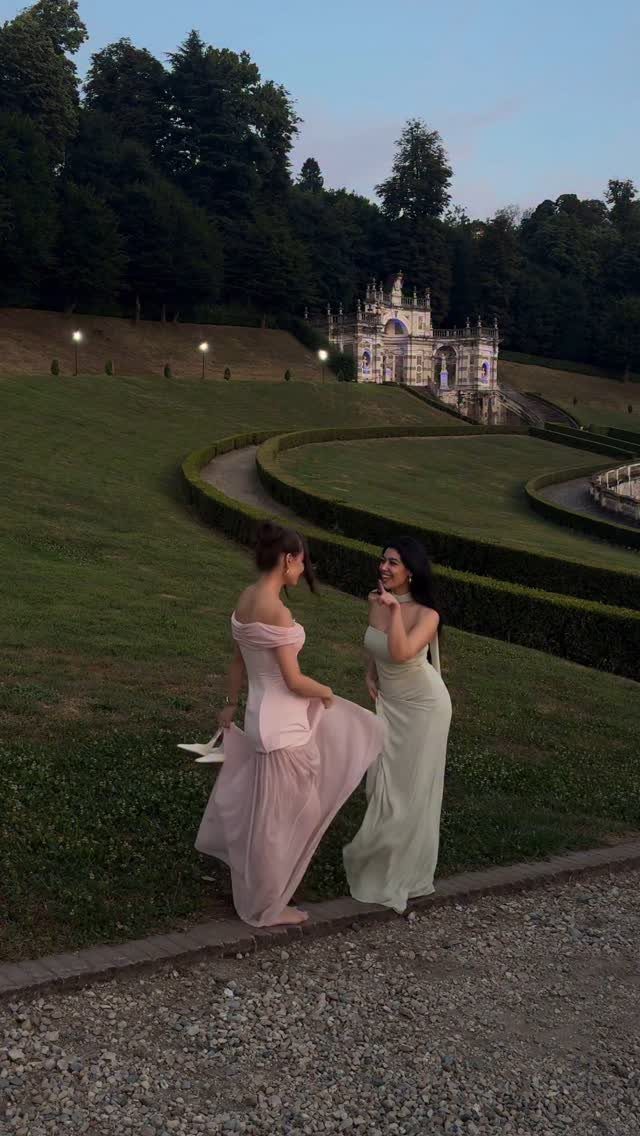 Two women in elegant long gowns walking through a manicured Italian garden at dusk.