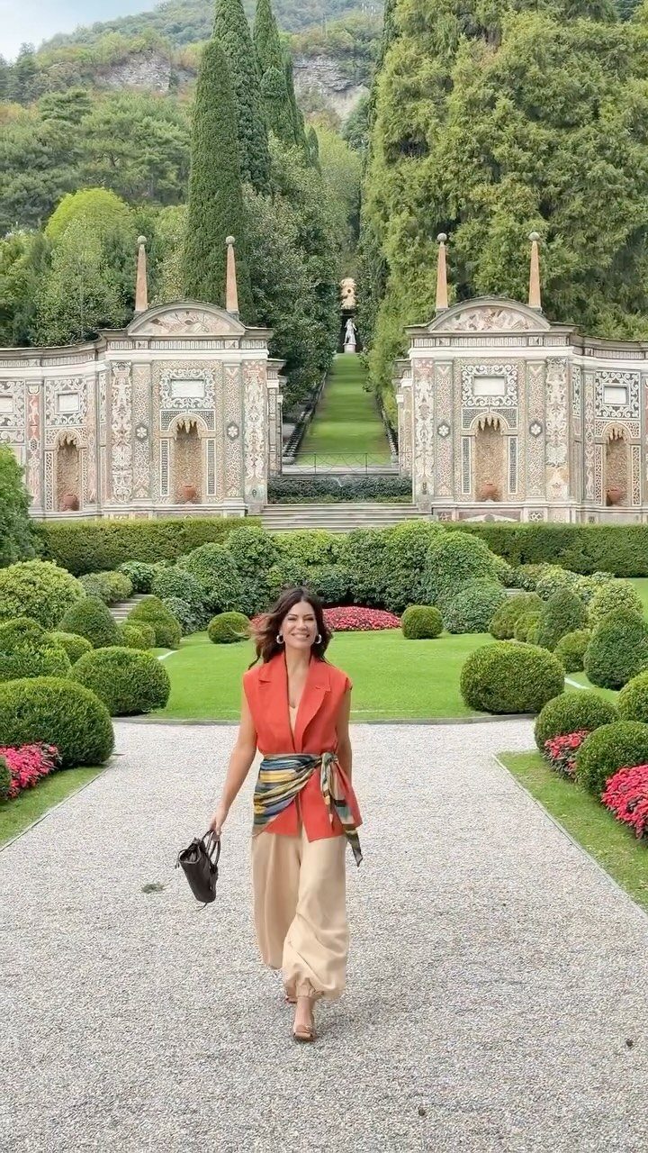 Woman in orange top and beige trousers walking through a manicured Italian garden path.