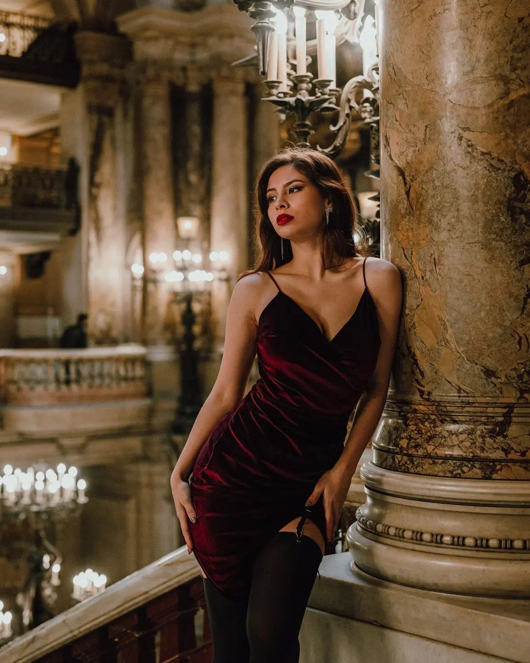 Woman in a burgundy velvet slip dress posing against a marble column in a grand hall.