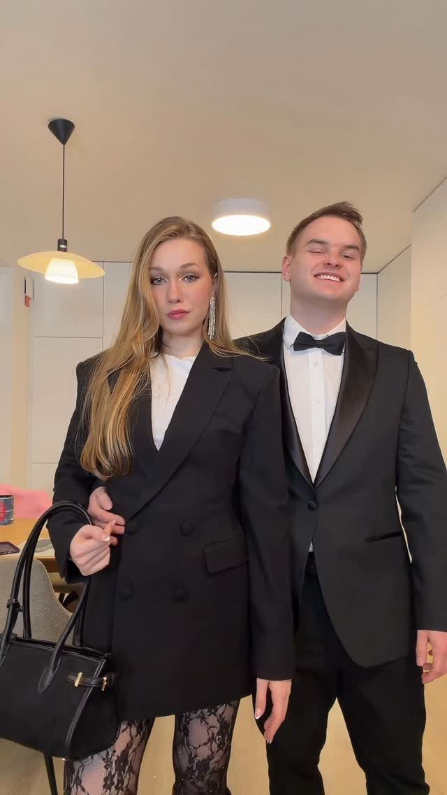 A man in a tuxedo and a woman in a black blazer posing together indoors.