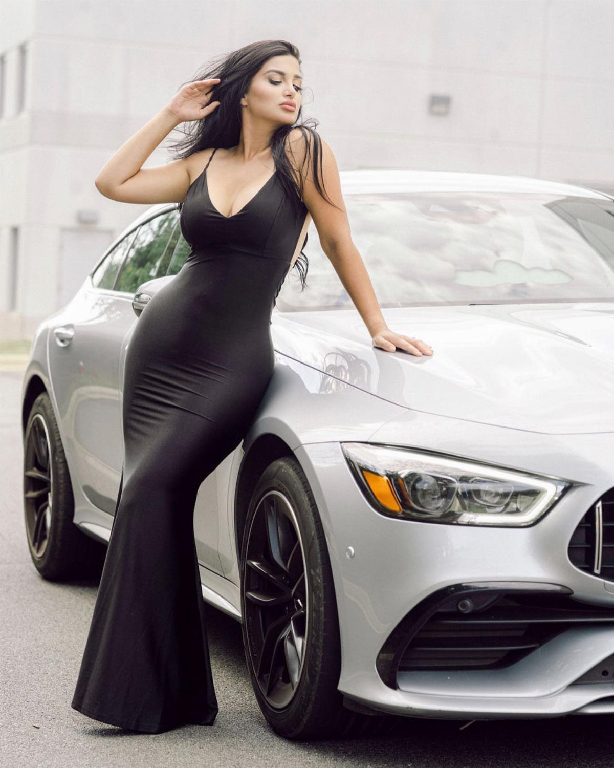 A woman in a long black evening gown leaning against a silver luxury car.