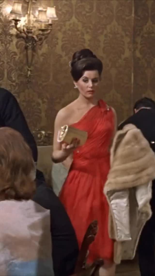 A woman in a red one-shoulder gown holding a gold clutch in a vintage room.