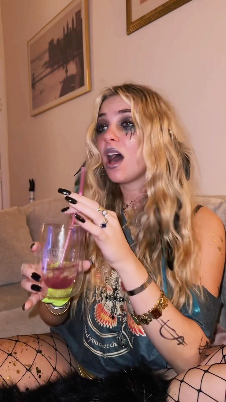 A blonde woman with dark smokey eye makeup and glittery tears holds a drink.