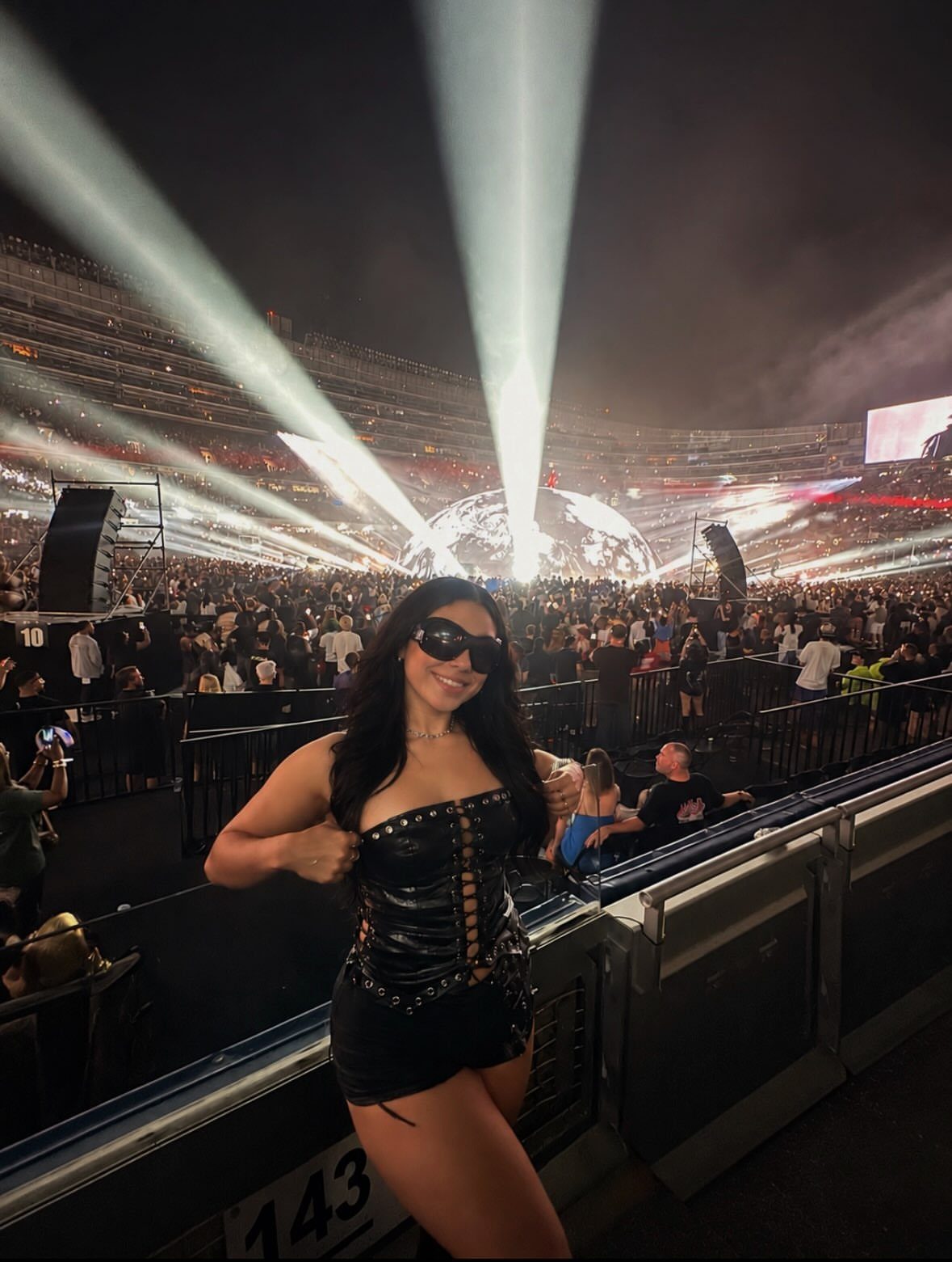 Woman in a black corset outfit posing at a large outdoor concert stadium at night.