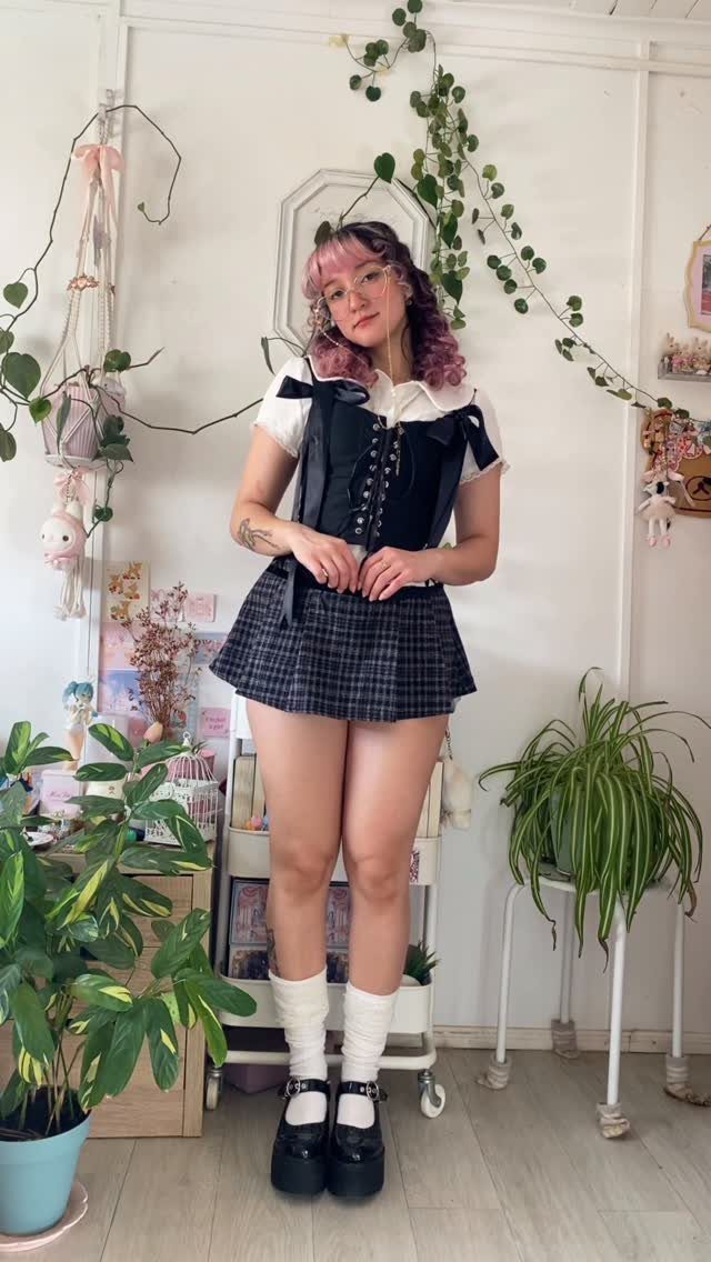 Woman with pink hair posing in a black corset, plaid skirt, and platform shoes.