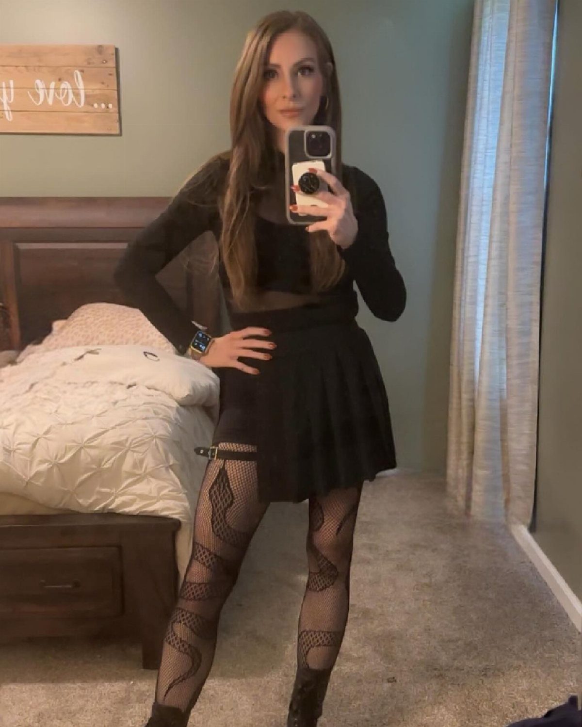 Woman in a black mesh top, short skirt, and patterned tights taking a mirror selfie.