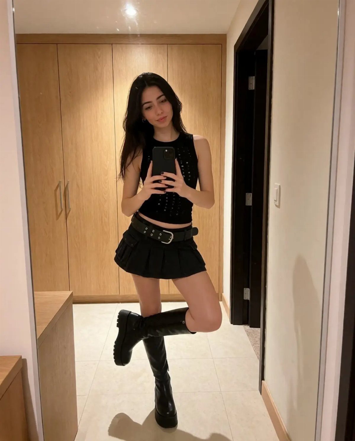 A woman posing in a black sleeveless top, pleated mini skirt, and black boots.