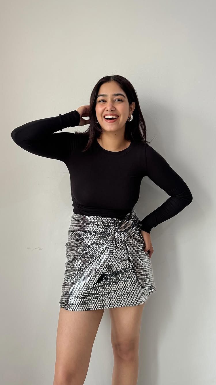 A smiling woman wearing a black long-sleeved top and a silver sequined mini skirt against a white wall.