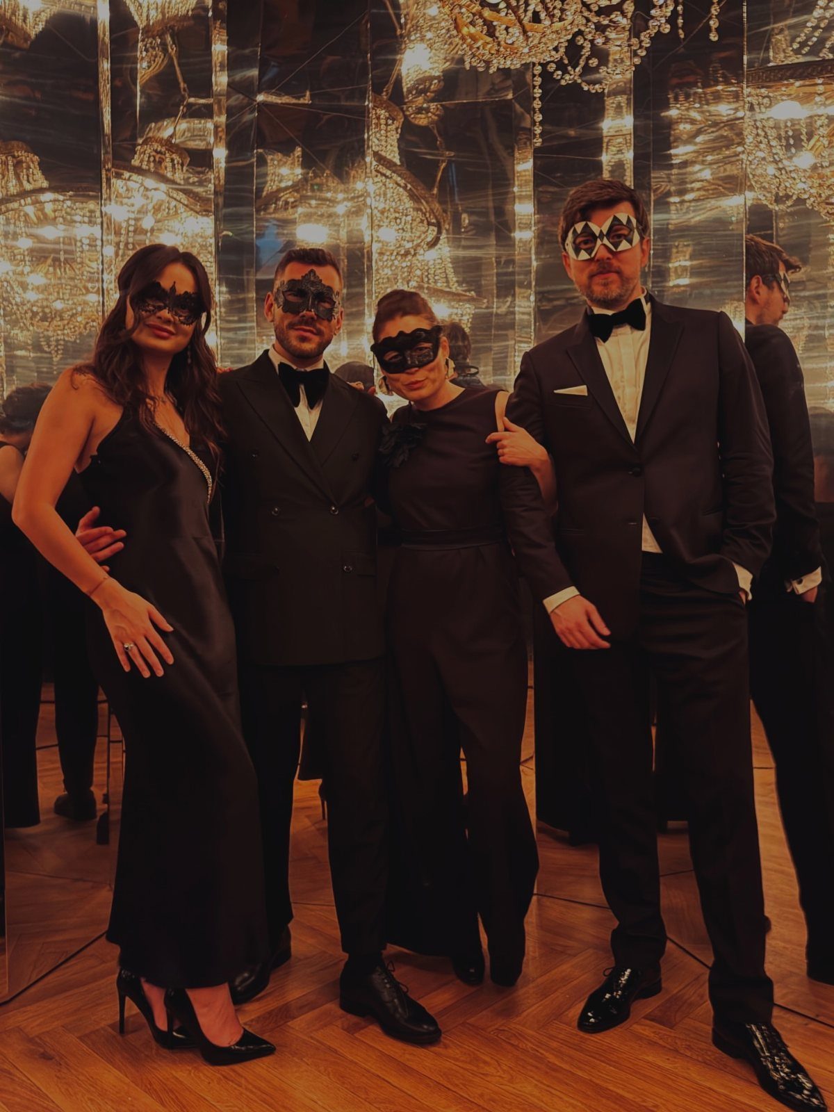 Four people in formal black clothing and decorative masks posing in a mirrored room.