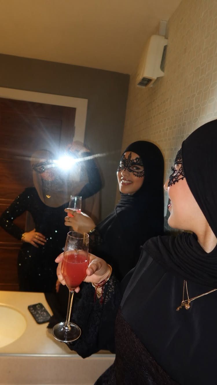Three women in black hijabs and lace masks posing for a mirror selfie with drinks.