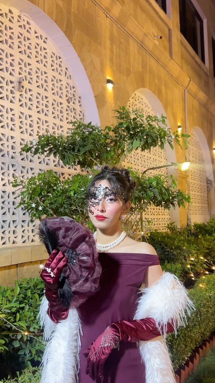 Woman in a burgundy dress and black lace mask holding a feathered fan.