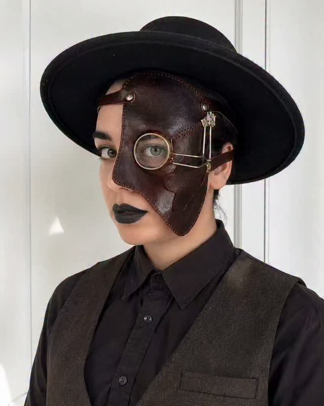 A person wearing a leather steampunk mask, a black hat, and a dark waistcoat.