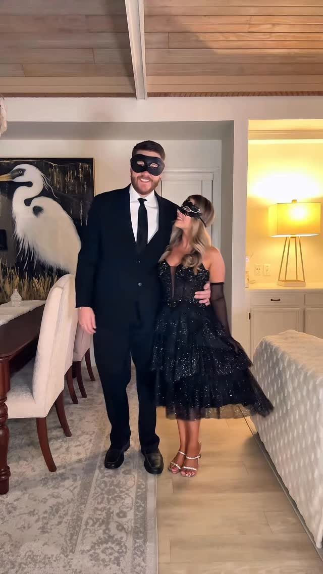 A man and woman wearing black masquerade masks and formal attire pose indoors.