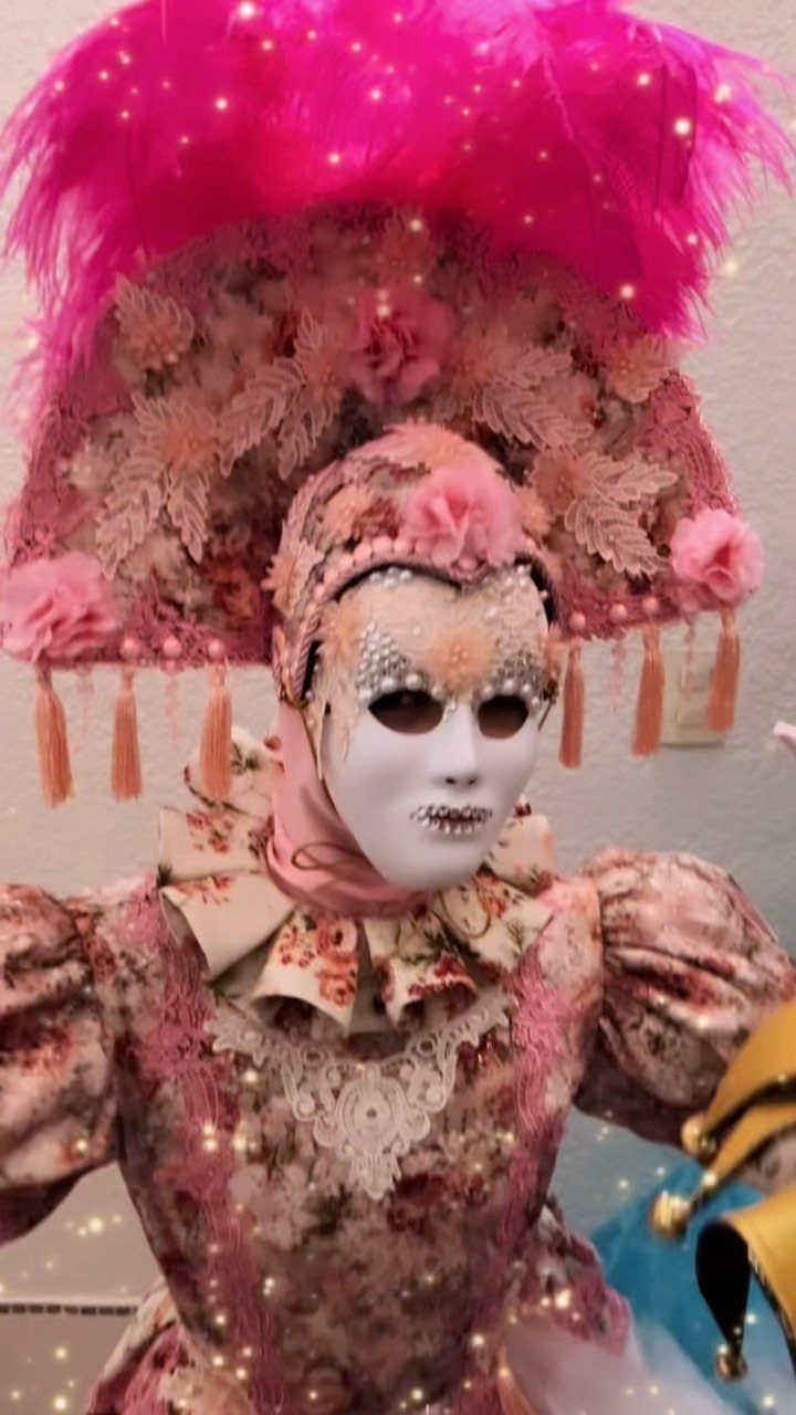 Person in an elaborate pink floral masquerade costume with a large feathered headpiece and white mask.