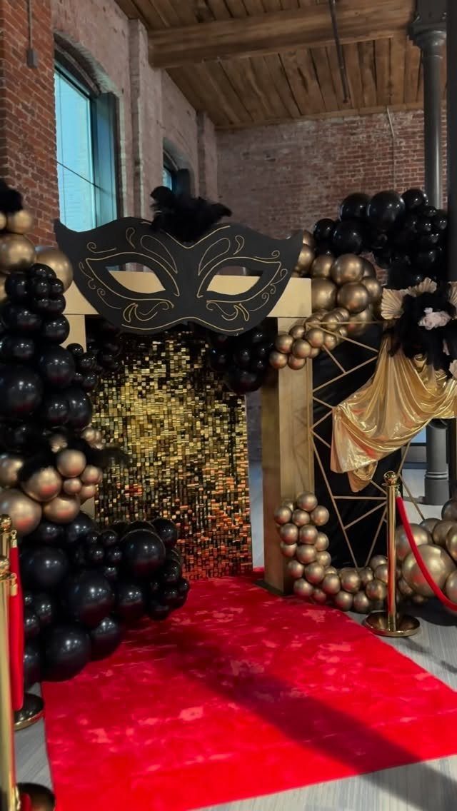 Masquerade event entrance with black mask, gold balloons, and a red carpet walkway.