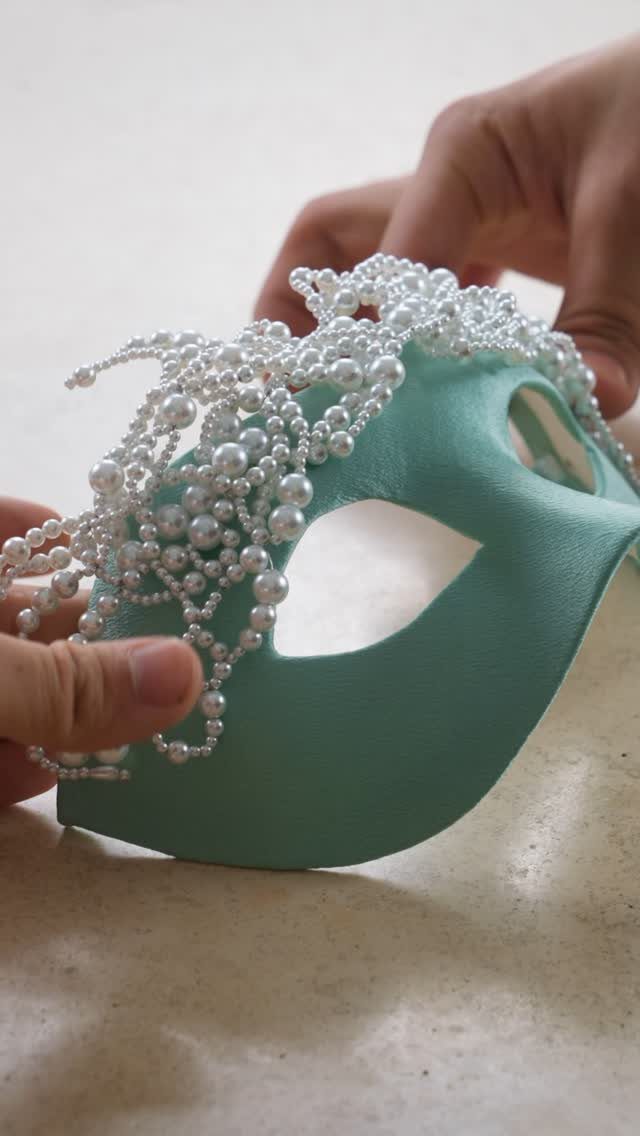Hands holding a teal masquerade mask adorned with many small white pearls.