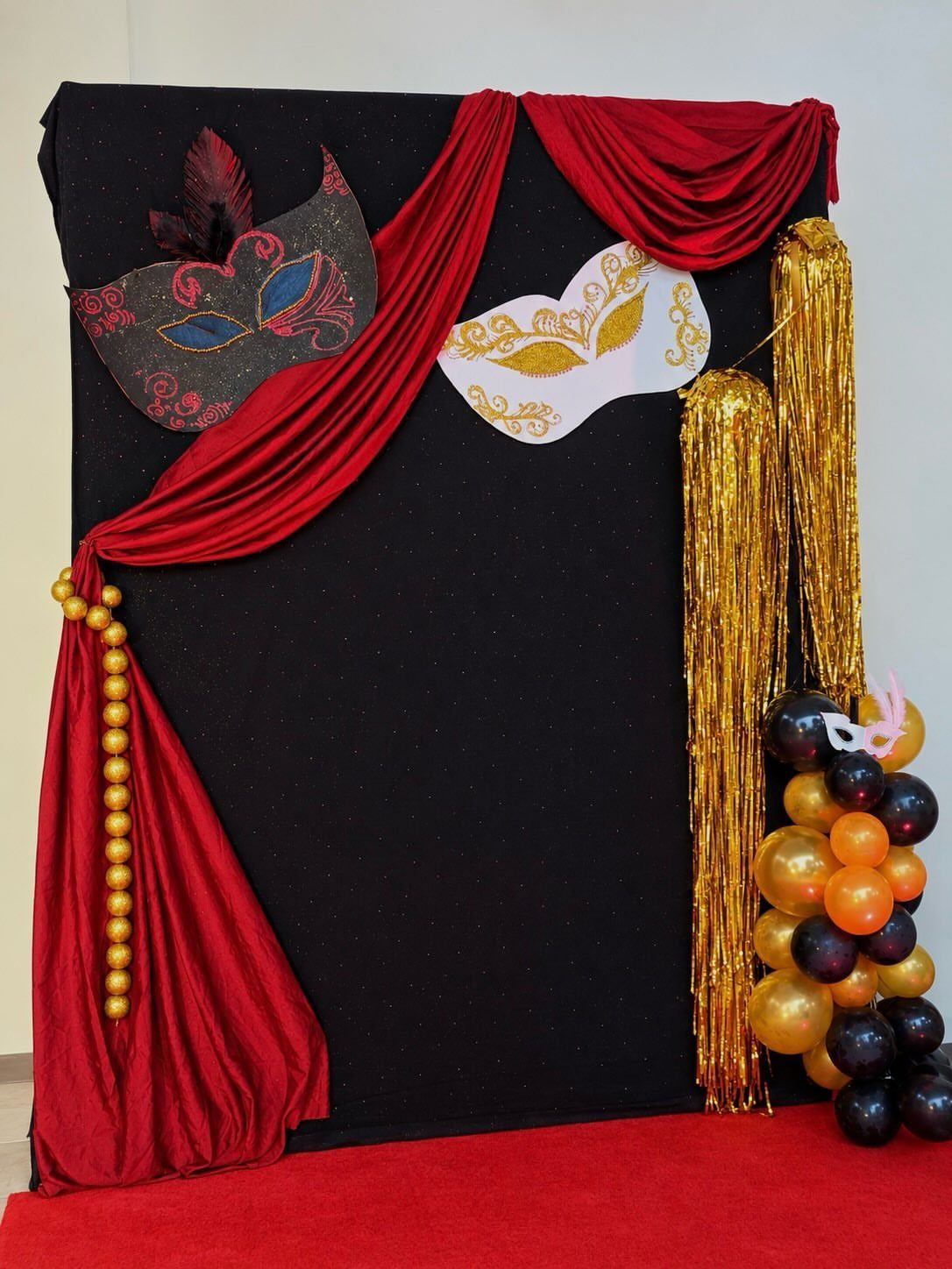A black masquerade themed photo booth backdrop with red drapes, masks, and gold balloons.