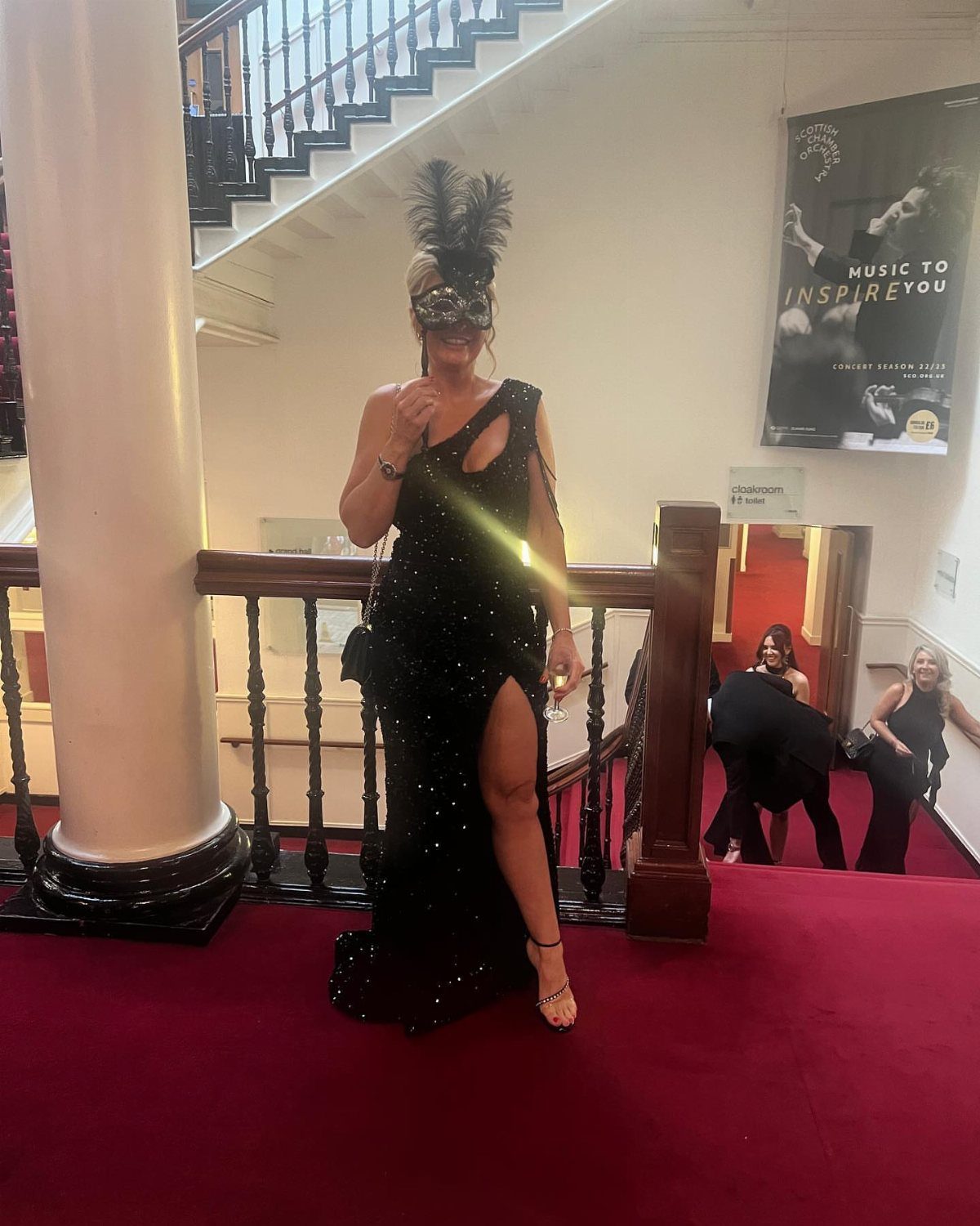 Woman in a black sequined gown and feathered mask standing on a red staircase.