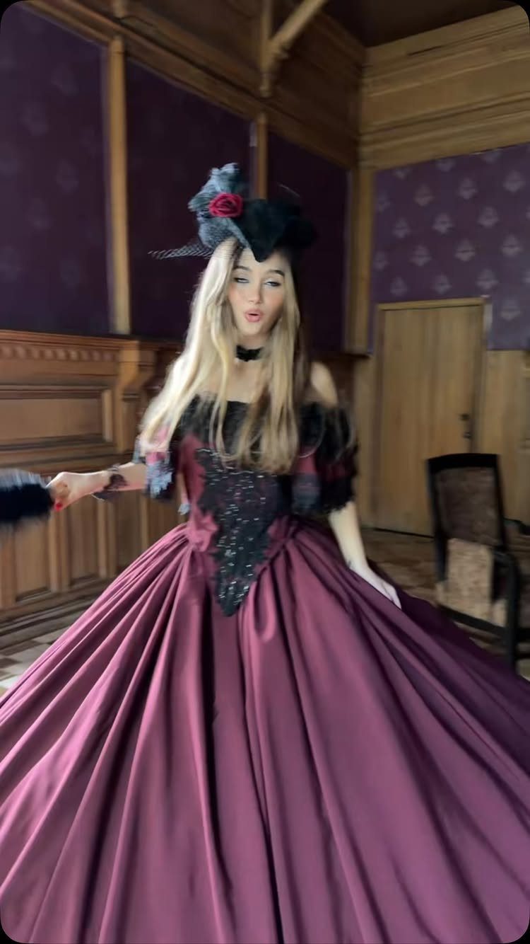 A woman in a voluminous burgundy ball gown and black fascinator posing in a room.