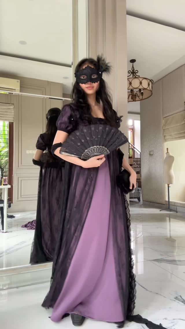 A woman in a purple gown and black lace mask holding a folding fan.