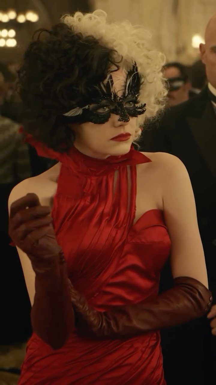 Woman in a red satin dress and black lace masquerade mask posing in a ballroom.