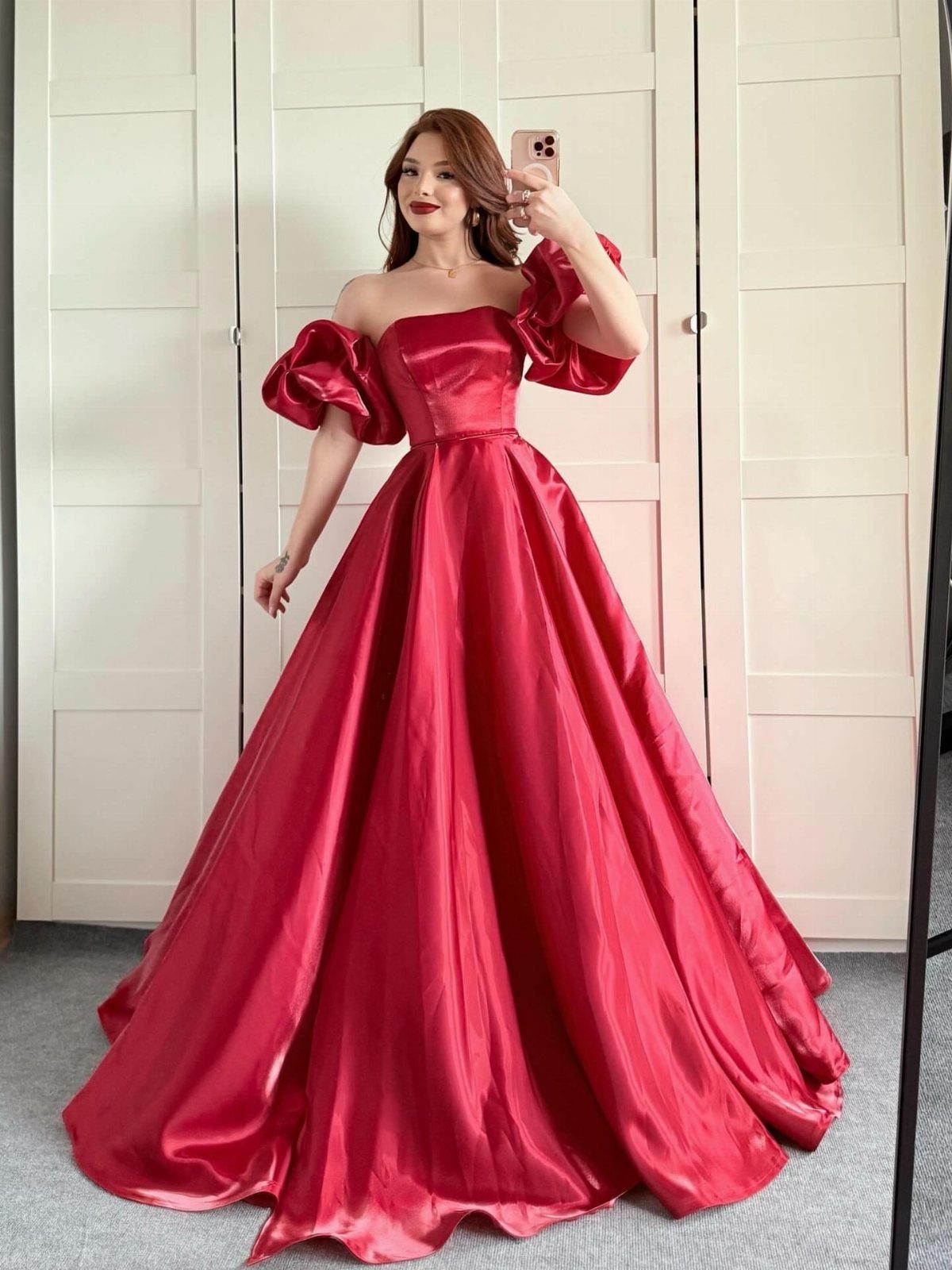 A woman wearing a voluminous red satin ball gown posing for a mirror selfie.