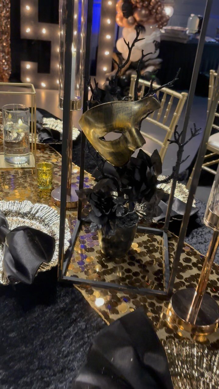 A gold masquerade mask sits in a black floral centerpiece on a decorated dinner table.
