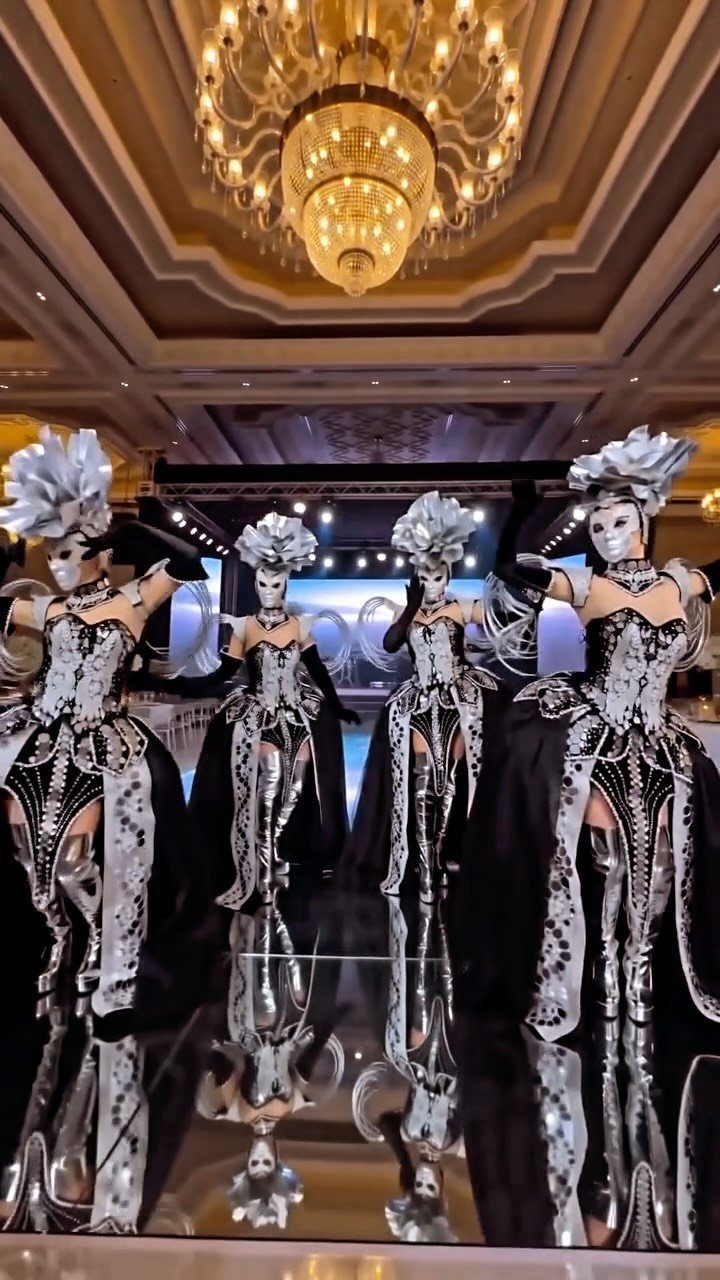 Four performers in silver and black masquerade costumes posing in a grand ballroom under a chandelier.