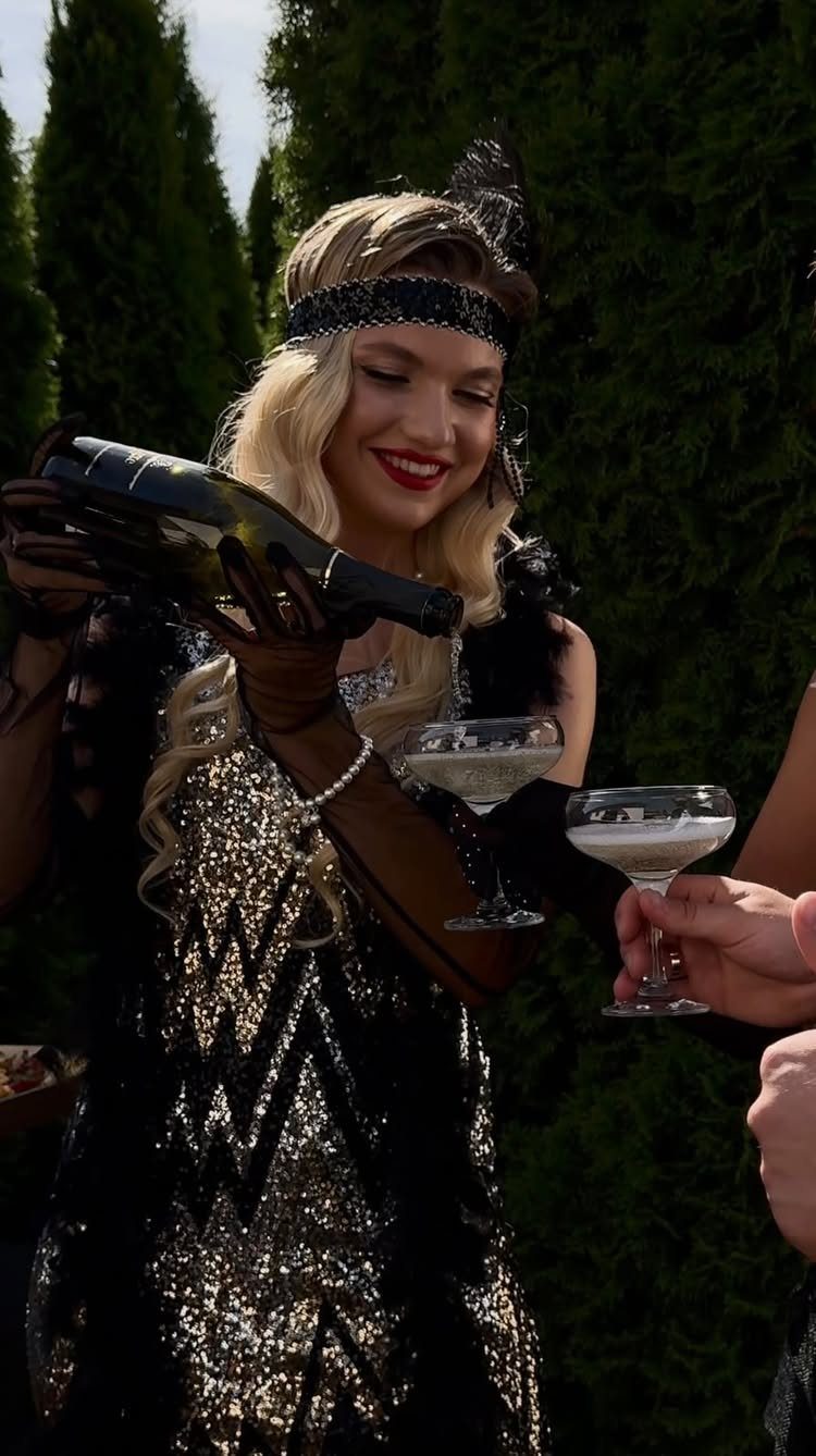 A woman in a black and gold sequined flapper dress pours champagne into a glass.