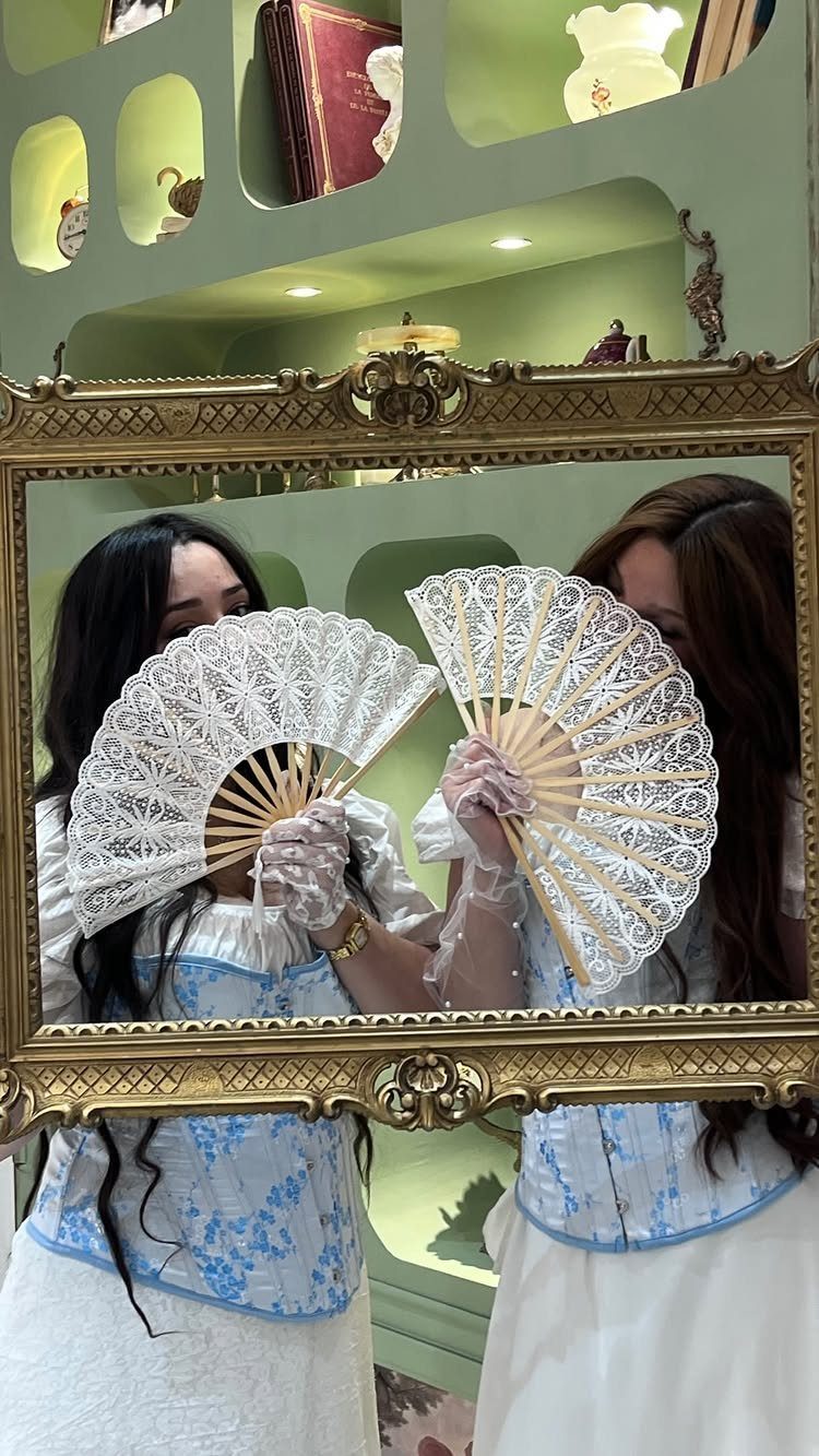 Woman in a blue floral dress holding a white lace fan in front of an ornate mirror.