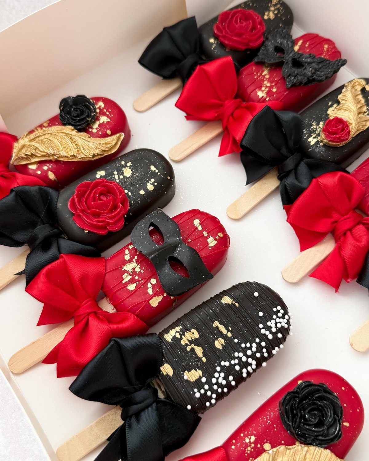 Decorated red and black cake pops with gold flakes, roses, and masks in a white box.