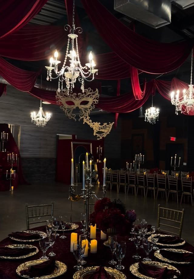 Formal dinner tables set with gold chargers and red napkins under hanging masquerade masks and chandeliers.