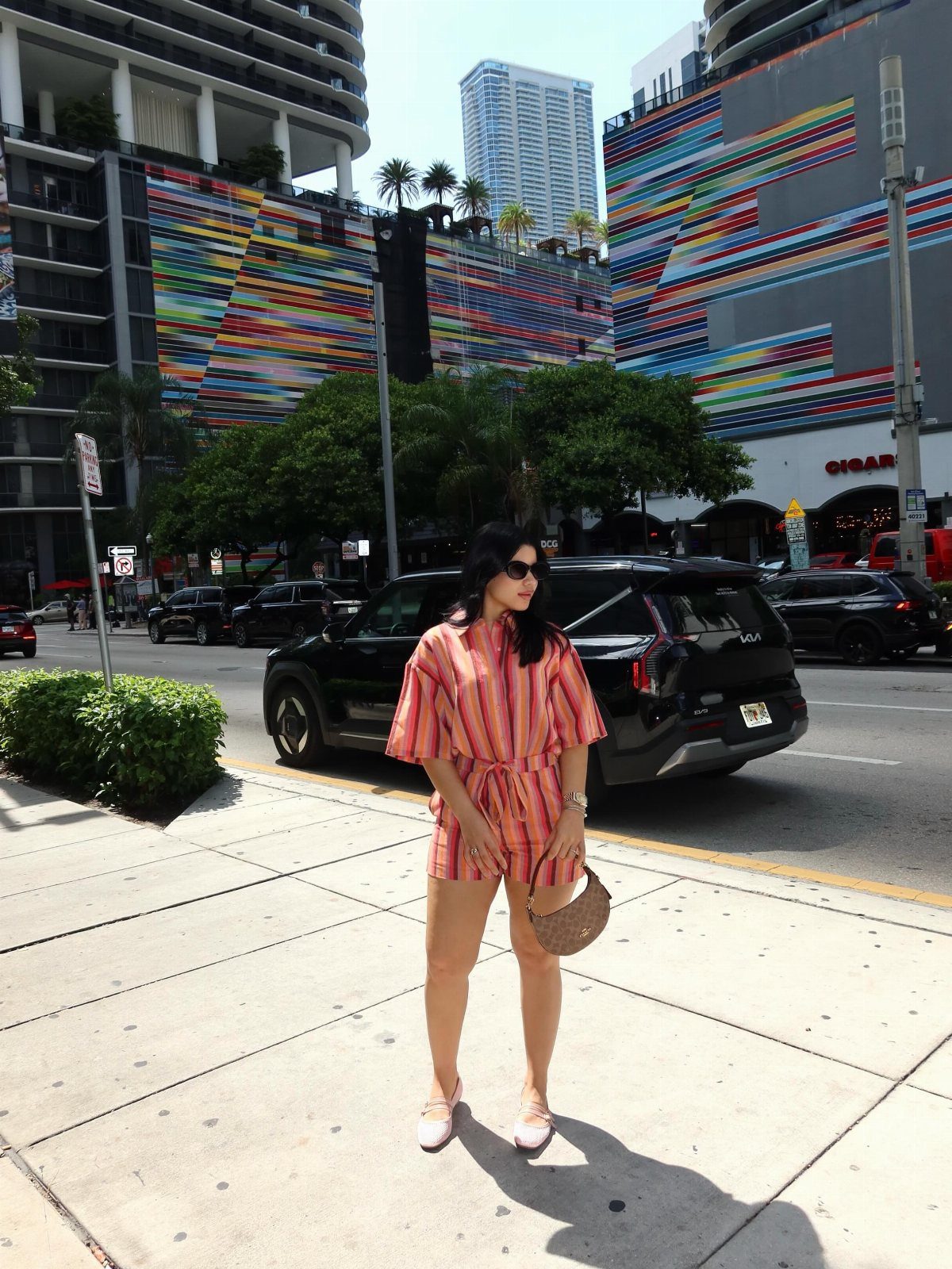 A woman in a pink striped breezy blouse and matching shorts standing on a city sidewalk.