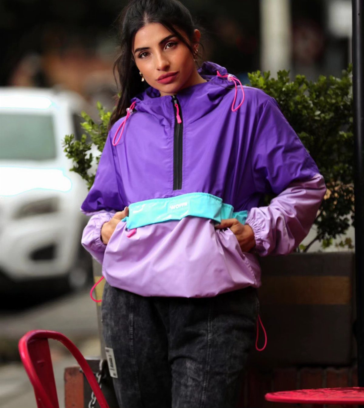 A woman wearing a colorful purple and pink windbreaker jacket standing outdoors near green plants.