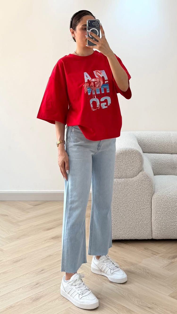 Woman in a red graphic t-shirt and light blue jeans posing for a mirror selfie.