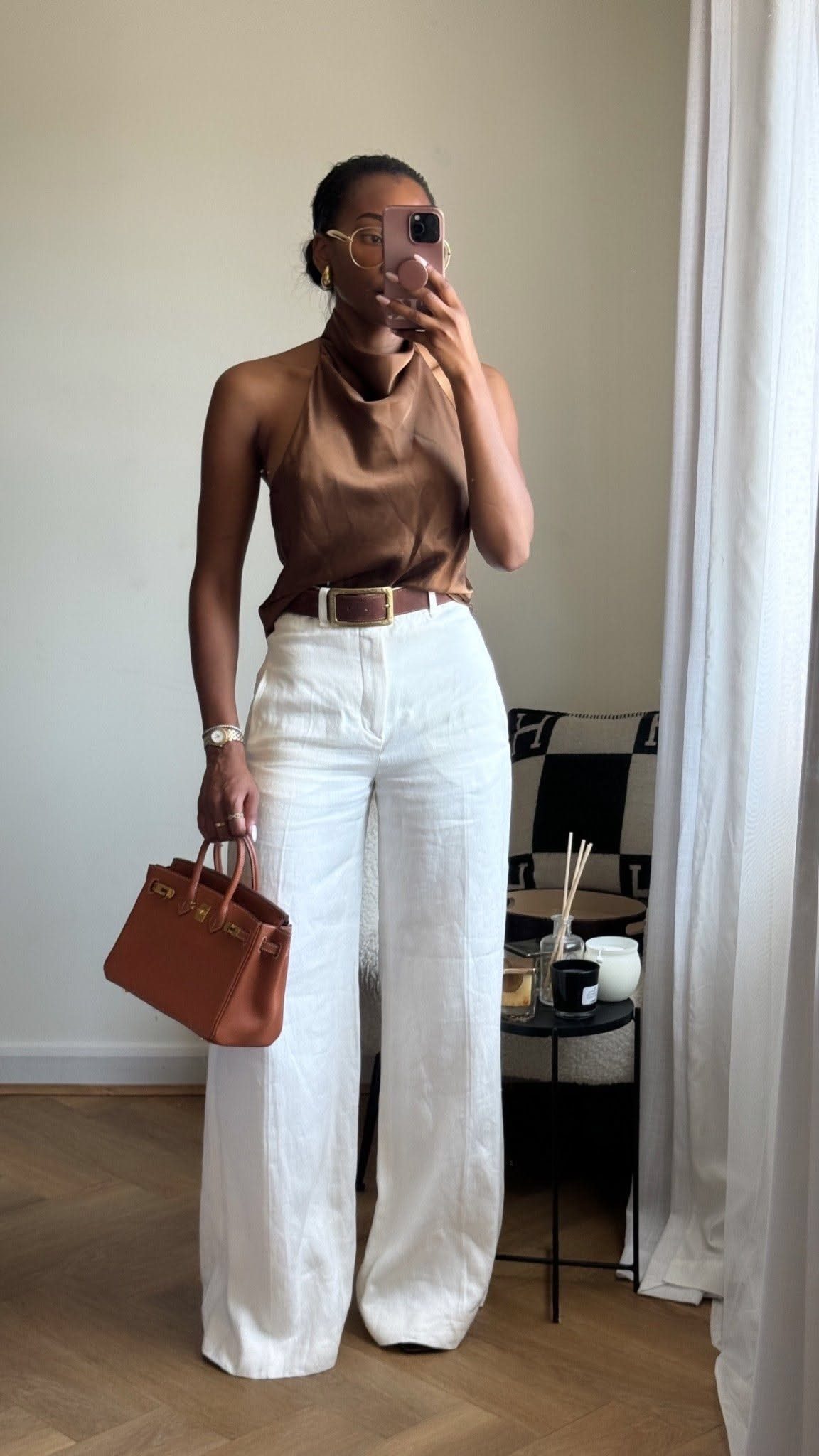 Woman wearing a brown silk halter top and white linen trousers holding a handbag.