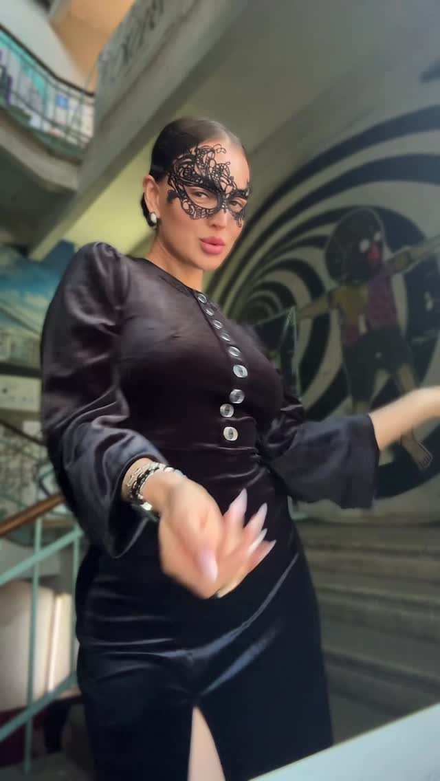 Woman in a black velvet dress and lace masquerade mask posing indoors.