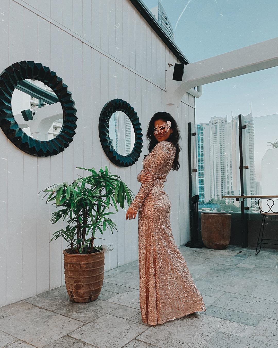 Woman in a rose gold sequined gown and mask posing on a rooftop terrace.
