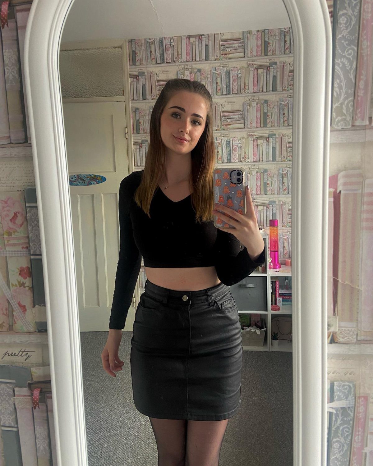 A young woman posing in a mirror wearing a black long-sleeved crop top and leather skirt.