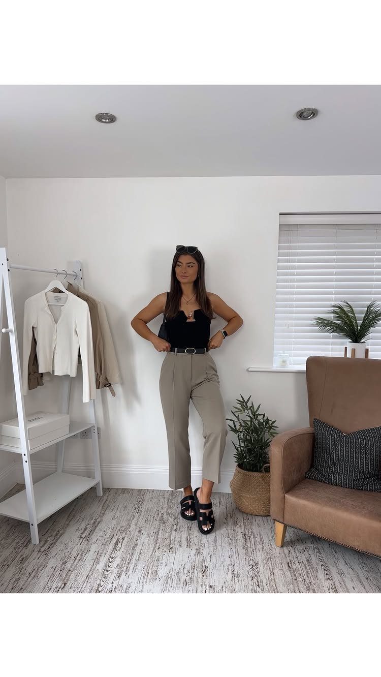 A woman posing in a black top, beige trousers, and black sandals in a bright room.