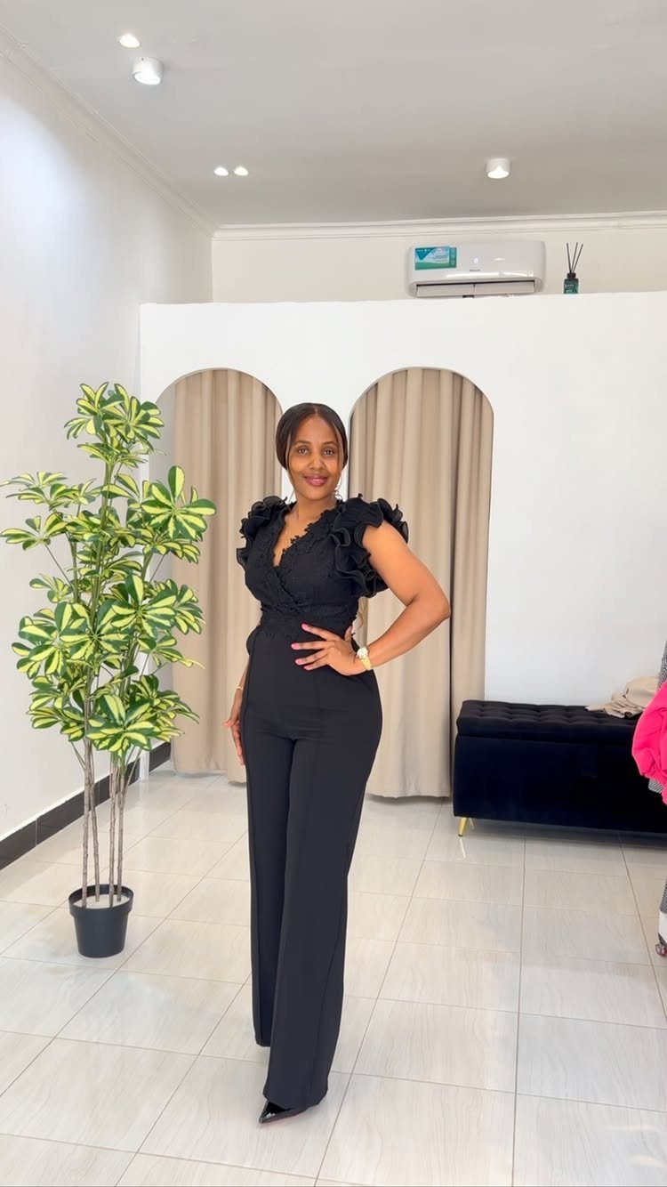 A woman wearing a black ruffled jumpsuit posing in a bright room with a plant.