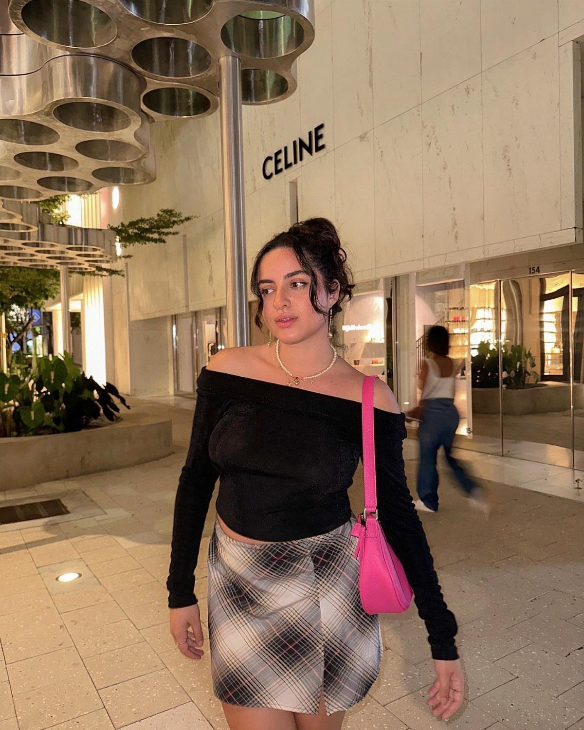 Woman in a black off-the-shoulder top and plaid skirt walking past a Celine store.