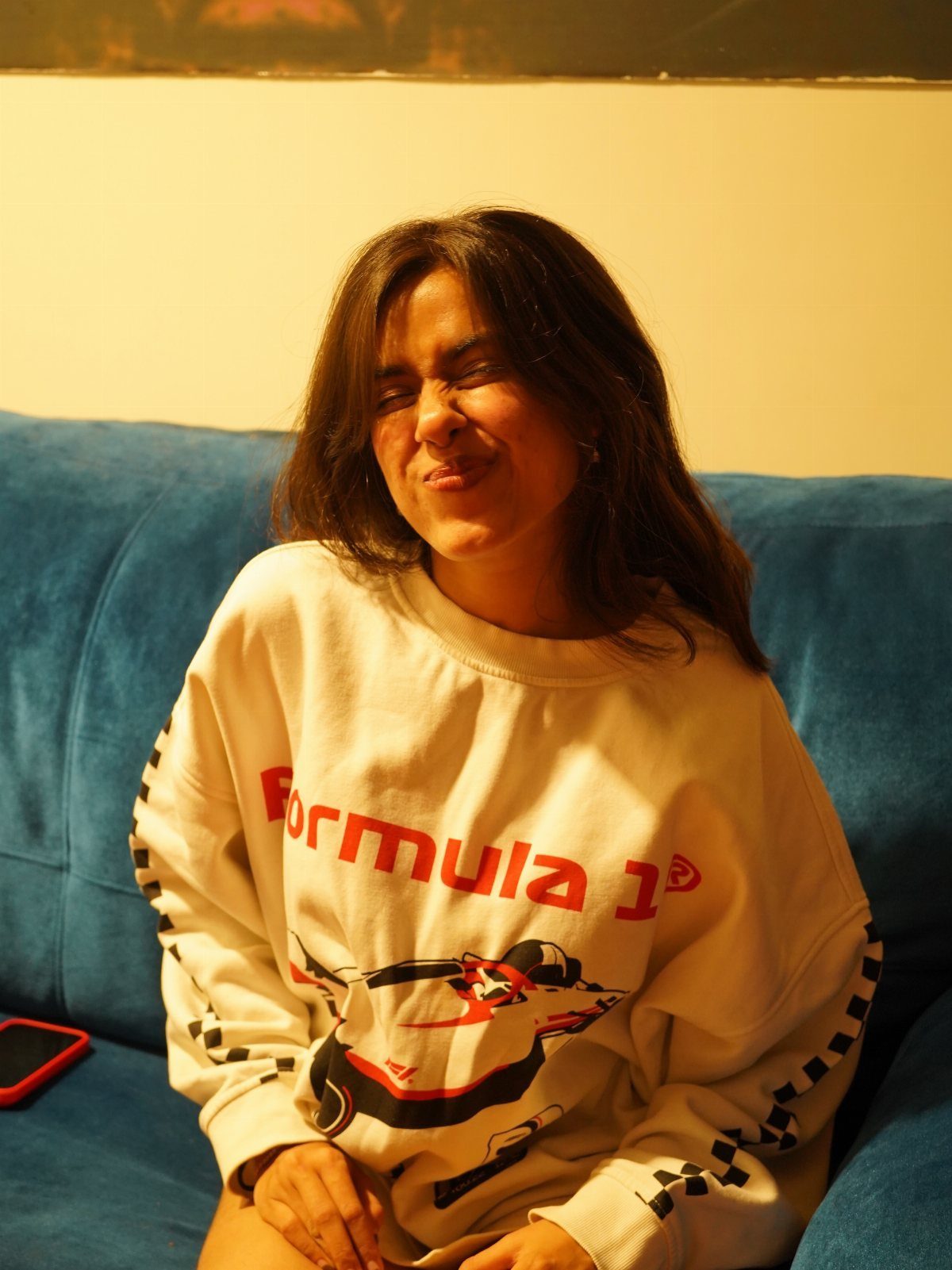 A woman sitting on a blue sofa wearing a white Formula 1 graphic sweatshirt.